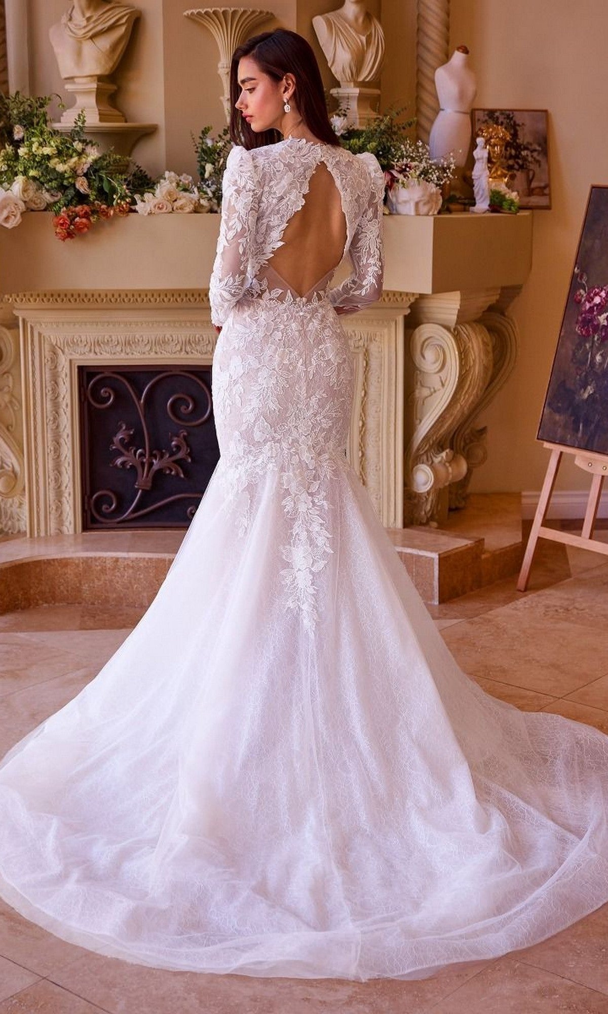 A woman stands indoors, facing away from the camera, wearing the Andrea and Leo WL036 long wedding dress—a lace mermaid gown with long sleeves, an open back, and a flowing train.